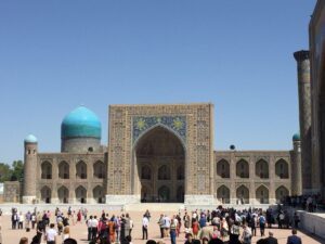 A large, ornate structure with a turquoise dome, intricate tile work, and a crowd of visitors in front under a clear blue sk…