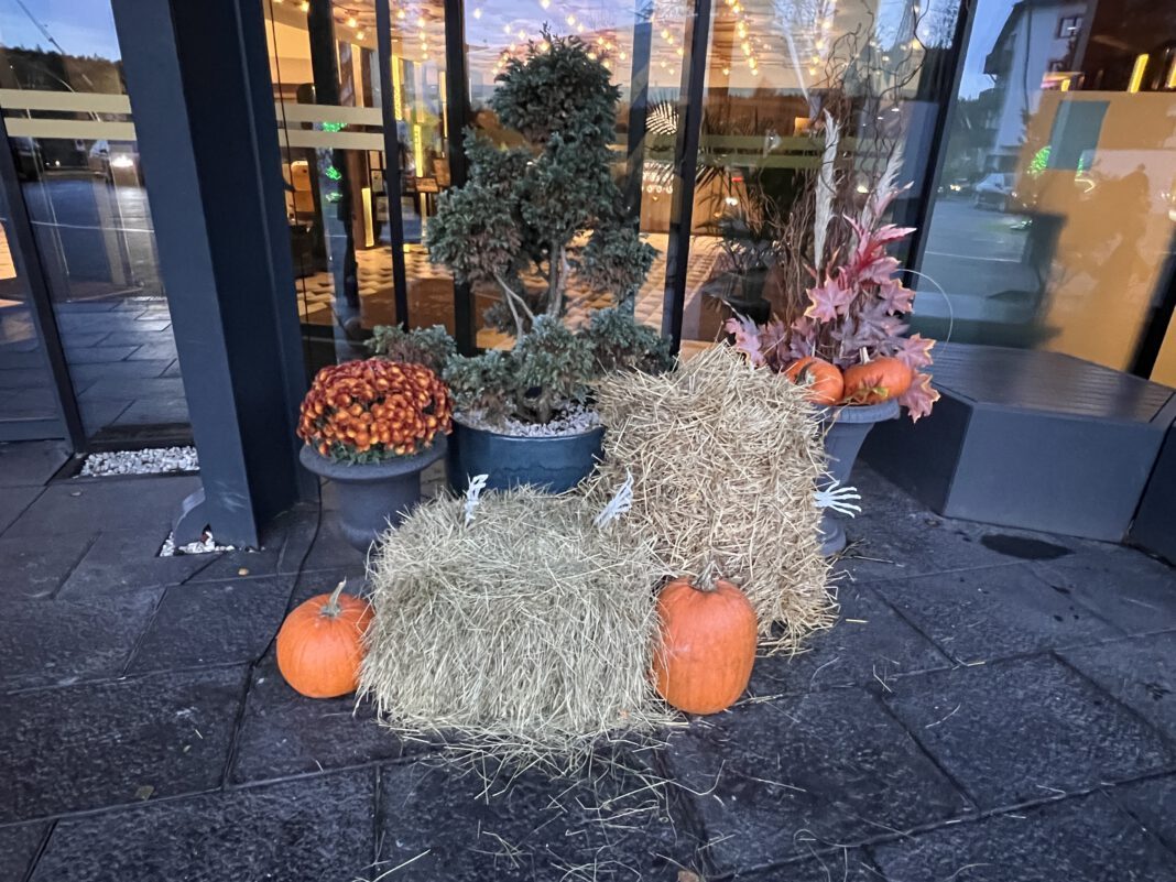 Decorative fall display featuring pumpkins, hay bales, and seasonal plants at a storefront entrance.